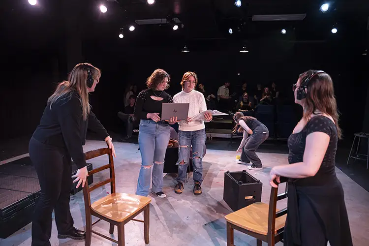 Theater rehearsal with people wearing headsets, arranging chairs and script on stage.