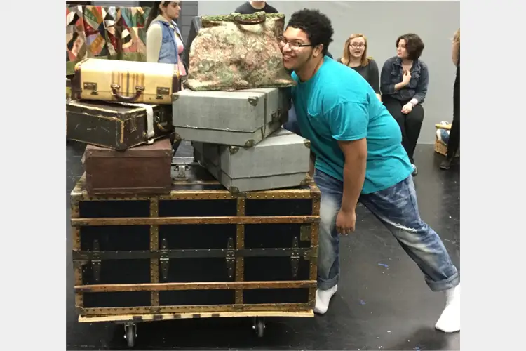 Person in a teal shirt leans against stacked suitcases on a rolling crate while others sit and stand in a studio space. Person in a teal shirt leans against stacked suitcases on a rolling crate while others sit and stand in a studio space.