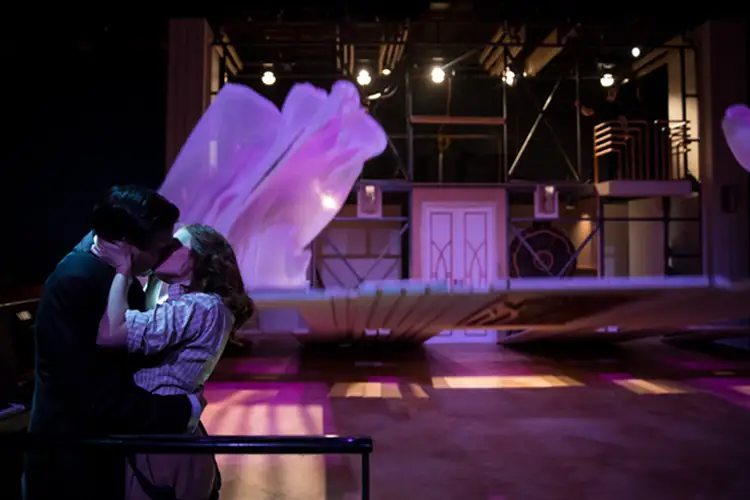 Two people sit close on a bench on a stage, leaning in and kissing, with a stylized set and soft purple lighting in the background. Two people sit close on a bench on a stage, leaning in and kissing, with a stylized set and soft purple lighting in the background.