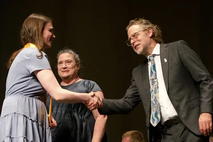 A student shakes hands with a professor on stage while a dean smiles in the background.
