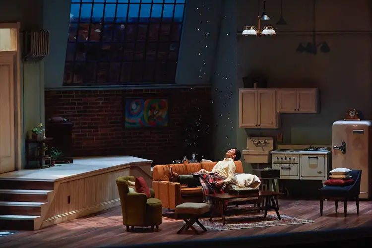 Stage set of an apartment: a person reclines on a couch under dim light, with kitchen and window backdrop and furniture arranged around a coffee table. Stage set of an apartment: a person reclines on a couch under dim light, with kitchen and window backdrop and furniture arranged around a coffee table.