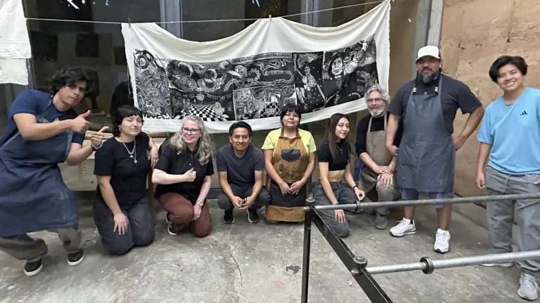 Group of artists pose in workshop before large black-and-white artwork on fabric.