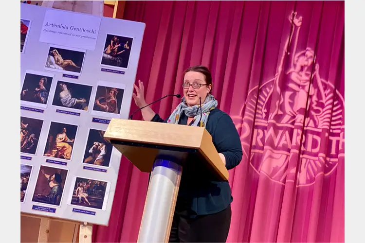 Speaker stands at a podium gesturing beside a display board of Artemisia Gentileschi paintings, with a red curtain backdrop and projected logo.