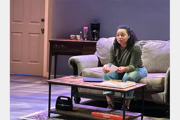 Person sits on couch holding papers behind a coffee table in a staged living room set with door, side table, and coffee maker. Person sits on couch holding papers behind a coffee table in a staged living room set with door, side table, and coffee maker.