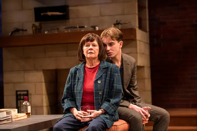Two performers sit close on a stage set with a fireplace, one holding a glass while the other leans in behind, both in modern attire. Two performers sit close on a stage set with a fireplace, one holding a glass while the other leans in behind, both in modern attire.