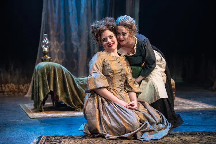 Two performers in period costumes on stage, one seated in a gold dress while another leans close; a table with lamp and draped cloth behind. Two performers in period costumes on stage, one seated in a gold dress while another leans close; a table with lamp and draped cloth behind.