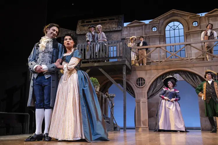Cast in period costumes perform on a theater stage with a balcony set; two figures stand foreground while others watch from above and behind. Cast in period costumes perform on a theater stage with a balcony set; two figures stand foreground while others watch from above and behind.