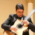 Leonela Alejandro in formal attire playing a classical guitar on a lit stage with pillars in background.