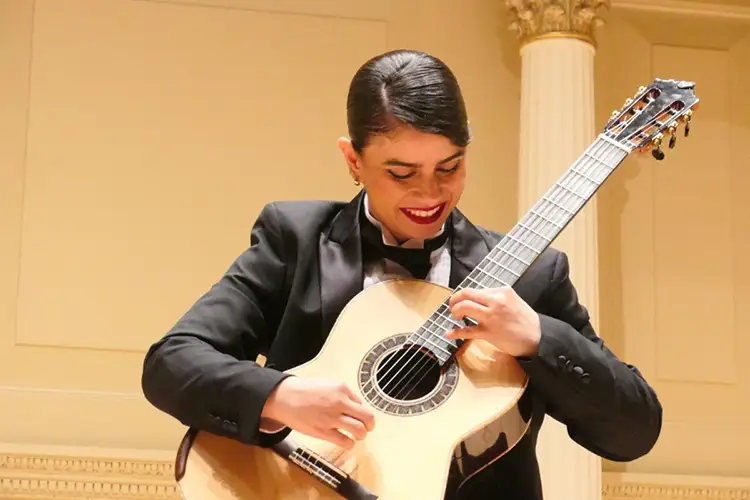 Leonela Alejandro in formal attire playing a classical guitar on a lit stage with pillars in background.