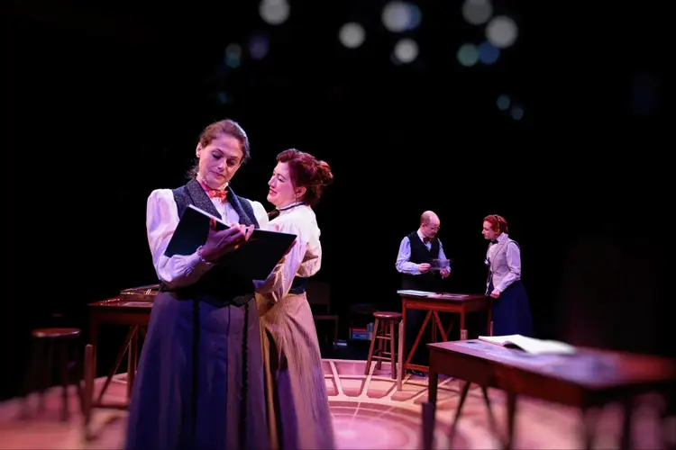 Actors on stage in period costumes; one writes in a book in the foreground while others stand by wooden tables under dim lighting. Actors on stage in period costumes; one writes in a book in the foreground while others stand by wooden tables under dim lighting.