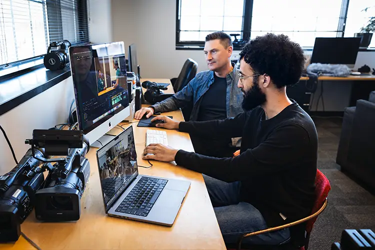 Film faculty and graduate student edit video footage at a workstation with professional cameras, monitors, and a laptop in a media production studio.