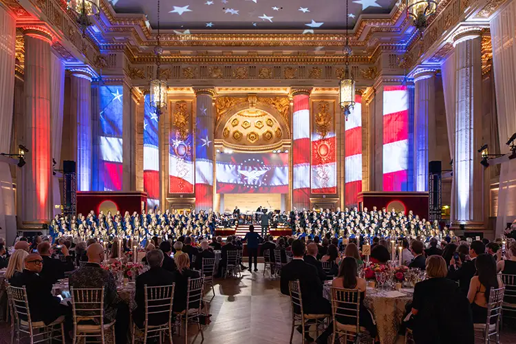 Large choir and orchestra perform before a seated audience in a grand hall with U.S. flag projections.