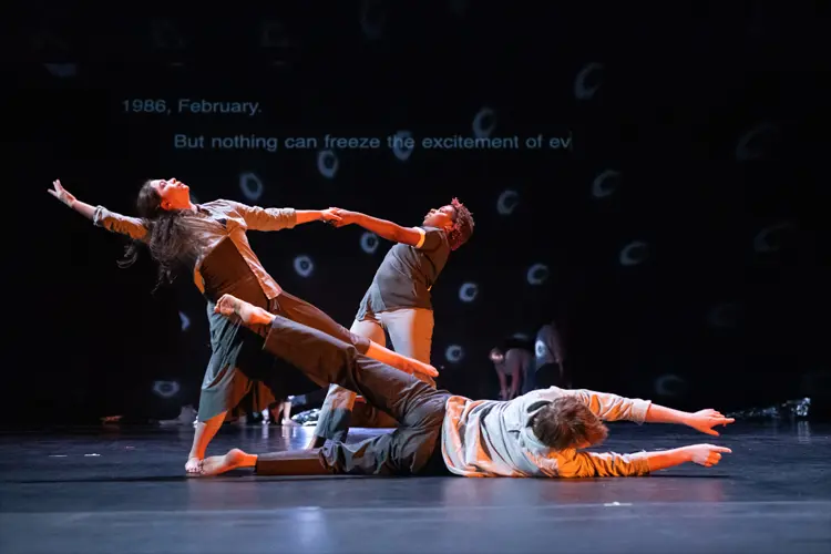 Three performers create a dynamic tableau onstage, with one reclining on the floor and two leaning back while linked by outstretched arms.
