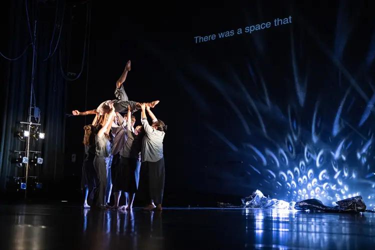 Group of performers lift a dancer overhead in a stage formation, with dramatic blue lighting and projected text in the background.