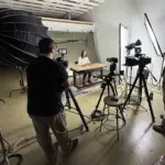 A film crew films a woman sitting at a desk in a well-lit space.