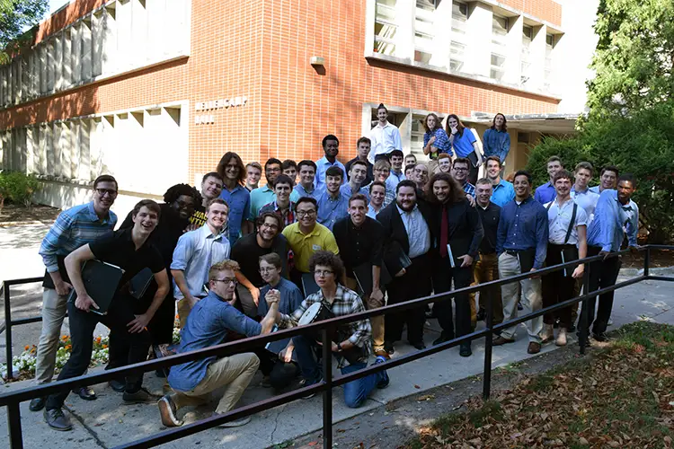 Large group gathered outside a brick academic building, posing together on a walkway before a performance. Large group gathered outside a brick academic building, posing together on a walkway before a performance.
