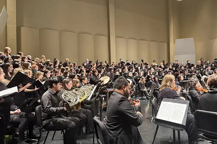 Large choir and orchestra perform together on a concert hall stage, with singers on risers and musicians in the foreground. Large choir and orchestra perform together on a concert hall stage, with singers on risers and musicians in the foreground.