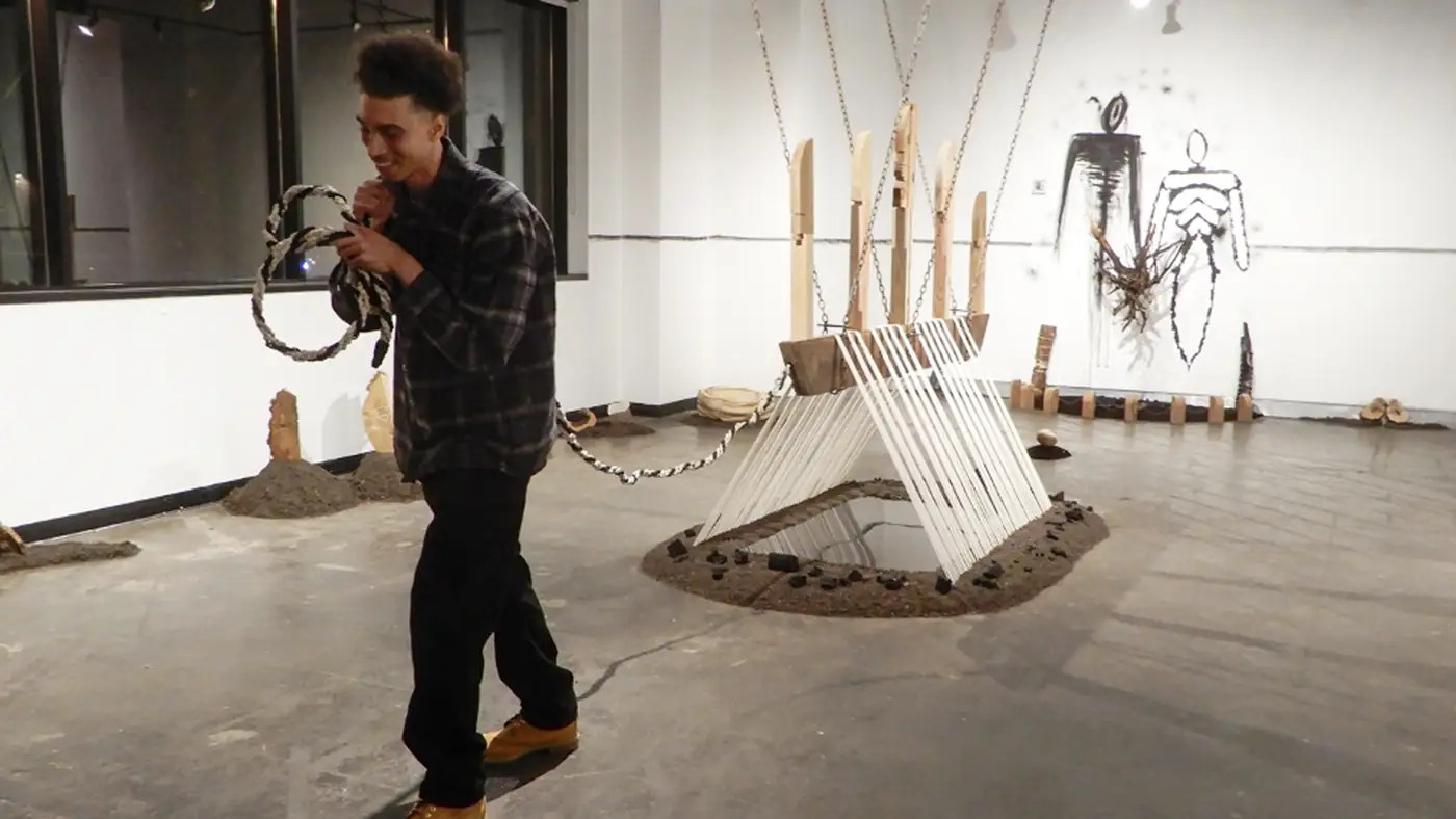 Person interacting with a large installation artwork featuring suspended structures, reflective surfaces, and wall drawings in a gallery space.