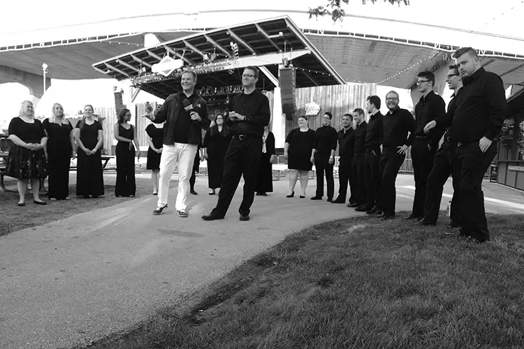 Choir members gather outdoors near a concert venue, listening as two speakers address the group before a performance. Choir members gather outdoors near a concert venue, listening as two speakers address the group before a performance.