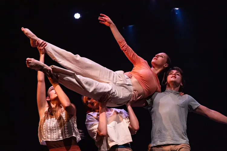 Performers lift a horizontal dancer overhead onstage, creating a balanced group formation under theatrical lighting