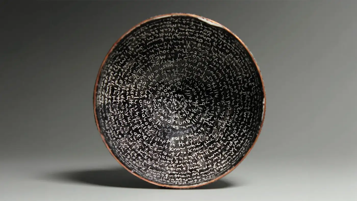 Black ceramic bowl lined with concentric rings of handwritten white text against a gray background.