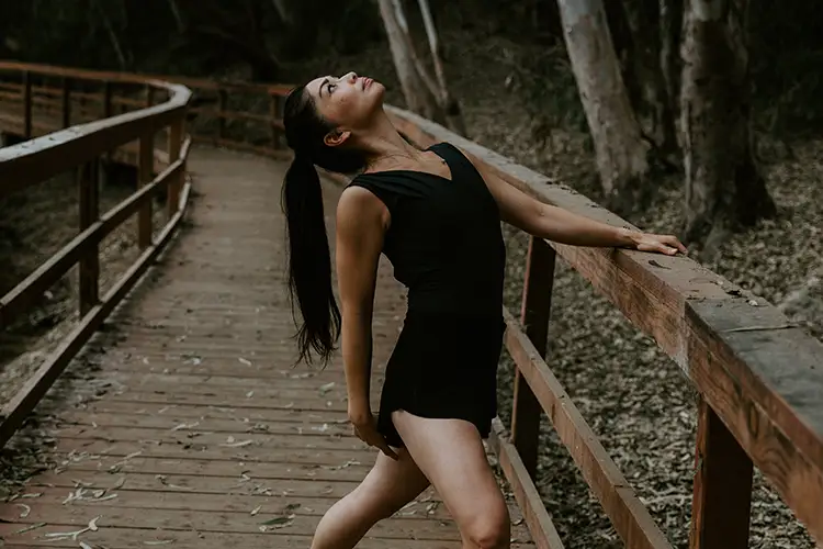 Person in black dancewear posing on a wooden boardwalk surrounded by trees.