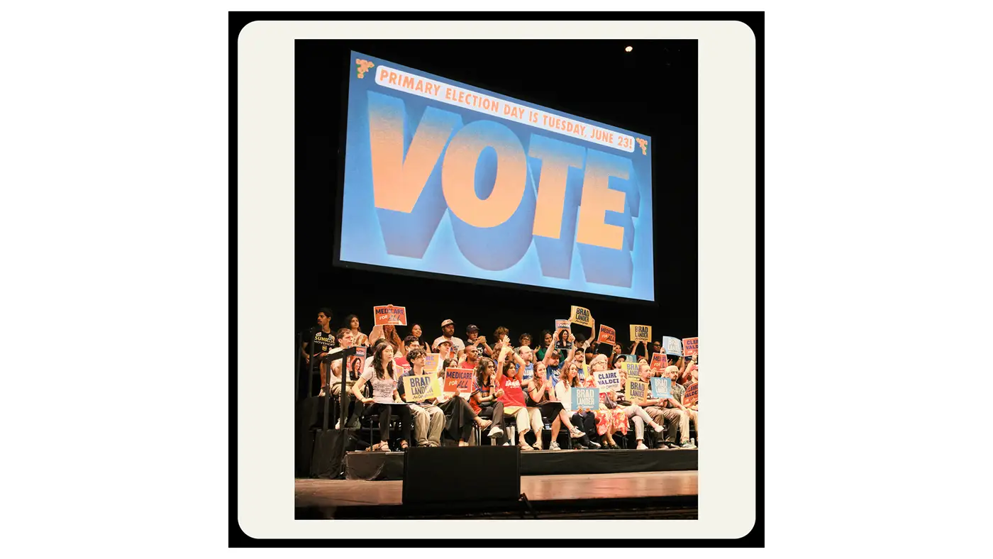 People holding campaign signs on a stage beneath a large “VOTE” projection promoting a primary election.