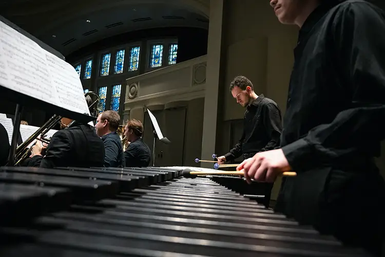 Musicians perform on percussion instruments in a concert hall, with a marimba in the foreground and stained-glass windows behind.