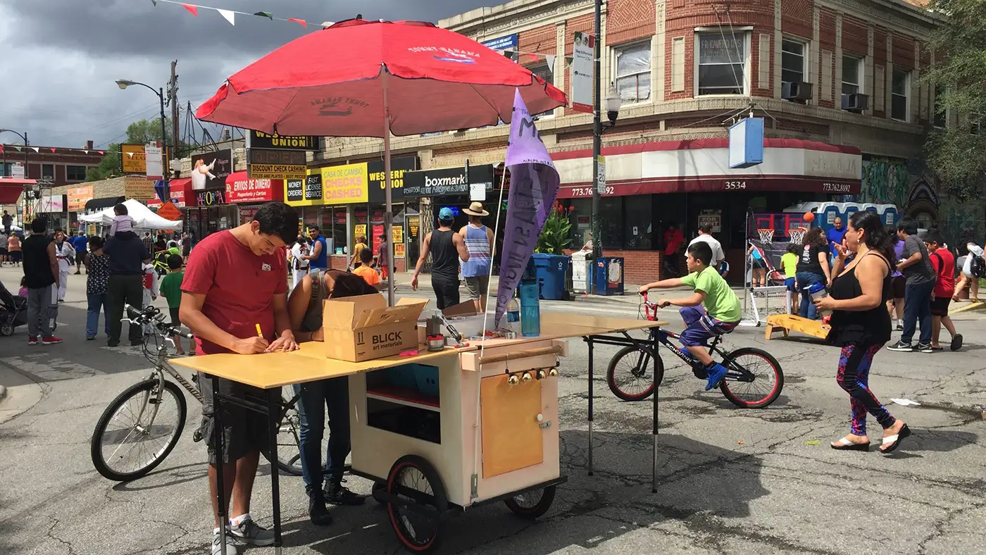 Community street festival with people gathering around a mobile art cart, bicycles, and neighborhood storefronts.