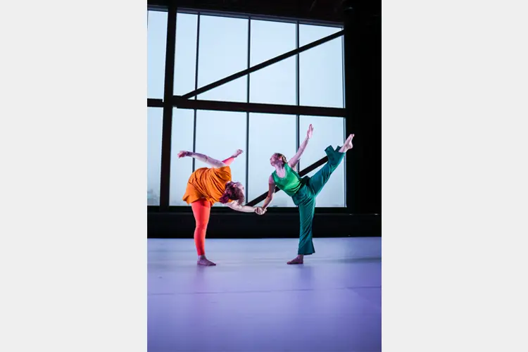 Two dancers balance in mirrored poses while holding hands on a stage, framed by a large geometric window backdrop.