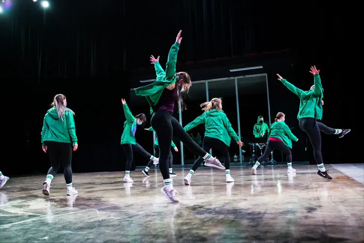 Group of dancers in matching green jackets move across a stage, arms raised and legs extended, with reflective panels in the background.