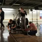 Film students adjust camera dolly tracks and lighting equipment in a spacious studio space.
