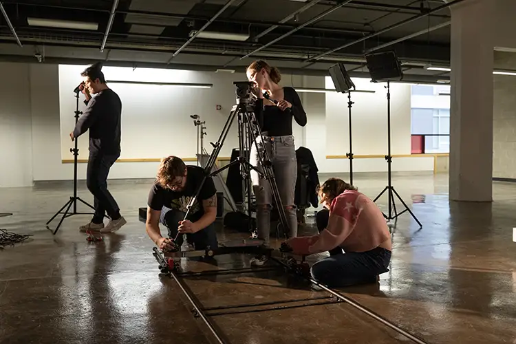 Film students adjust camera dolly tracks and lighting equipment in a spacious studio space.
