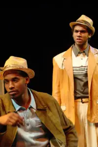 Two performers in straw hats and vintage clothing stand on a stage under warm lighting.