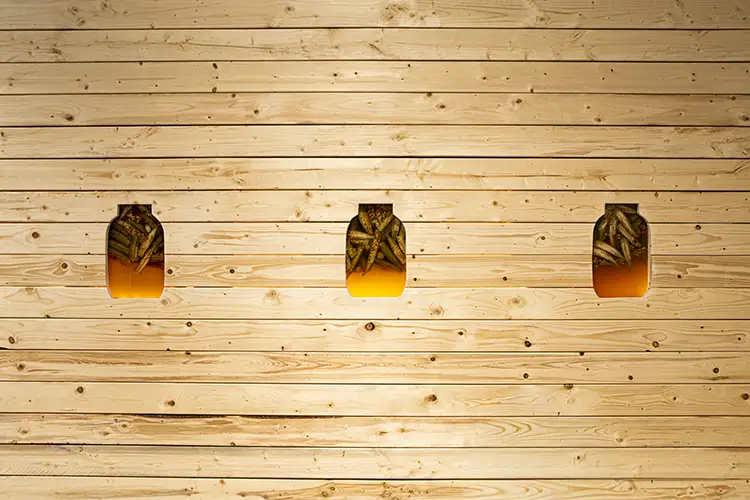 Jars of pickled okra behind a blonde wooden wall with jar shaped cut-outs Jars of pickled okra behind a blonde wooden wall with jar shaped cut-outs