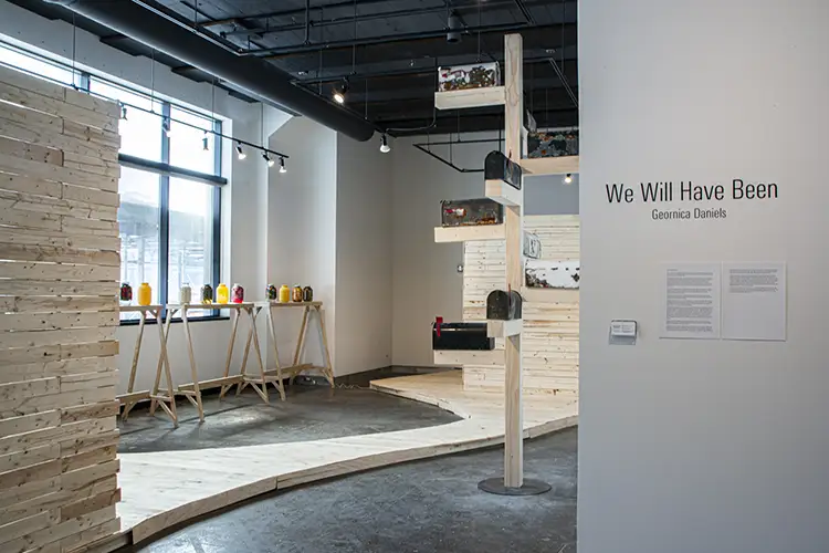 A gallery room with a wall reading "We Will Have Been, Geornica Daniels" with a wooden path leading between jars of pickled okra and mailboxes on a stand A gallery room with a wall reading "We Will Have Been, Geornica Daniels" with a wooden path leading between jars of pickled okra and mailboxes on a stand
