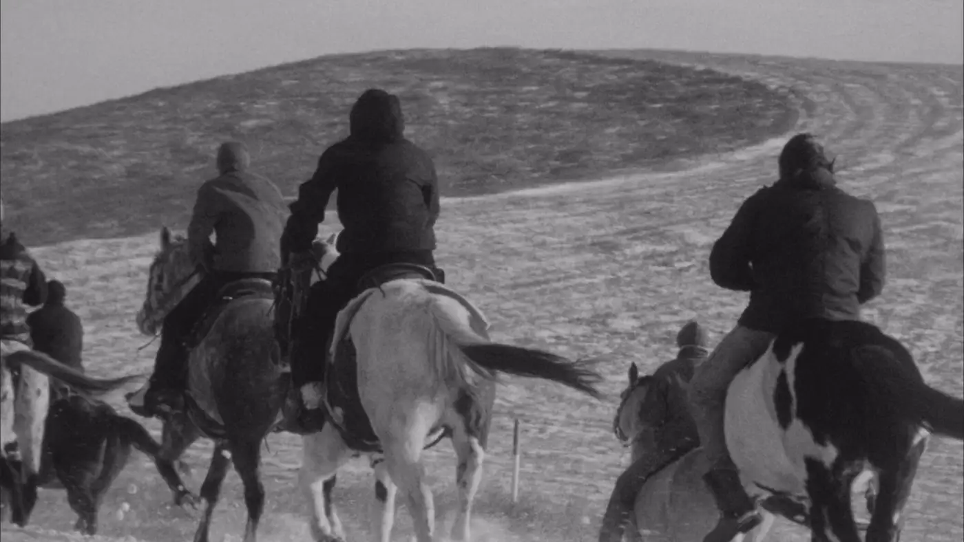 Riders on horseback travel across a snowy hillside in a black-and-white landscape, viewed from behind along a winding trail.