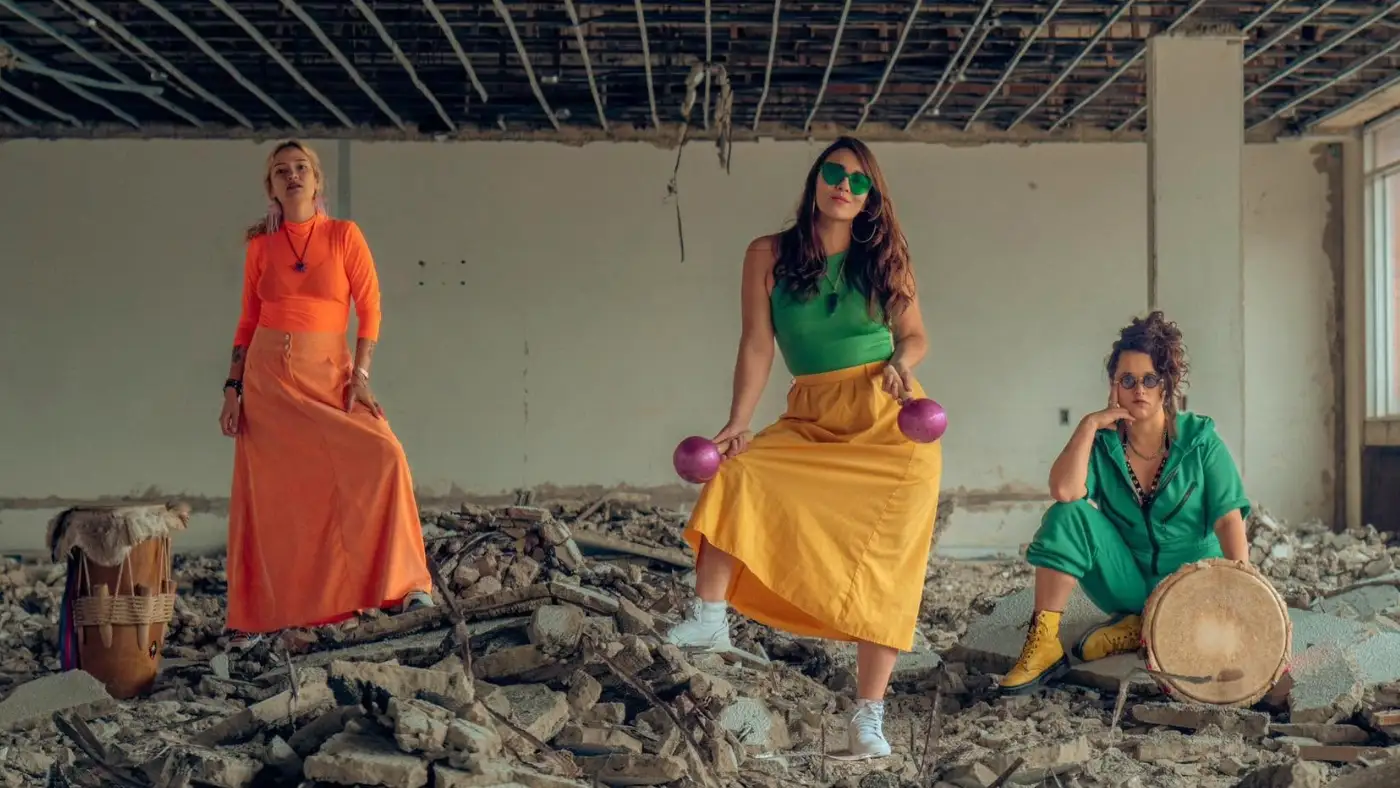 Three performers in bright orange, green, and yellow outfits pose among rubble with hand drums in a stripped interior space.