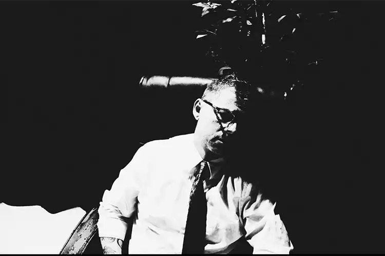 A man in a button-up shirt and tie sits in a chair. The image is black and white.