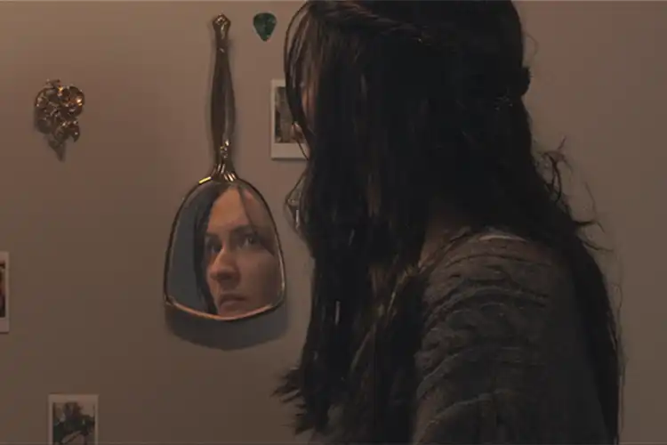 A young woman stares into a mirror as she walks down a hallway covered with photos and trinkets.