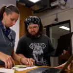 Three people gather around a table in a studio, examining a printed design while using open laptops and discussing a project.