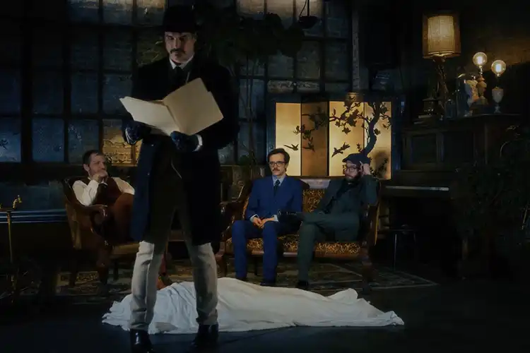 A mustached man in a nice suit reads a news paper with his back turned to three other dapper men who are sitting on a couch.