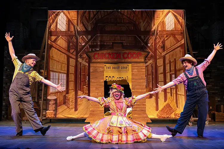 Kaegi does the middle splits in a frilly orange dress. She is in front of a barnyard-themed set and surrounded by two of her male co-stars.