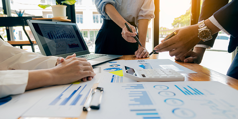 Business professionals reviewing charts and graphs on a table, with a laptop, calculator, and printed reports during a meeting.
