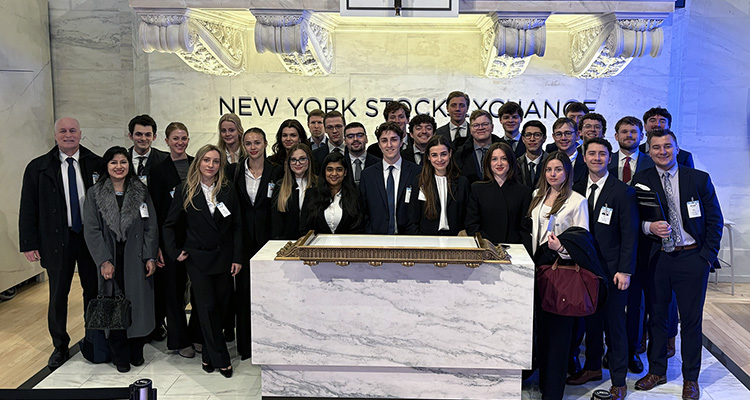 Large group of college students in professional suits standing in front of a New York Stock Exchange sign.