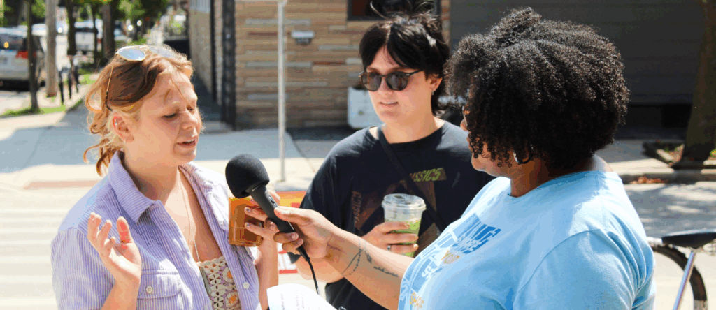 A Story Fellow with C21's Story Cart program holds a microphone up to the mouth of a young woman who is talking and gesticulating. Another person looks on from the side. They are standing on a tree-lined sidewalk on a sunny day.