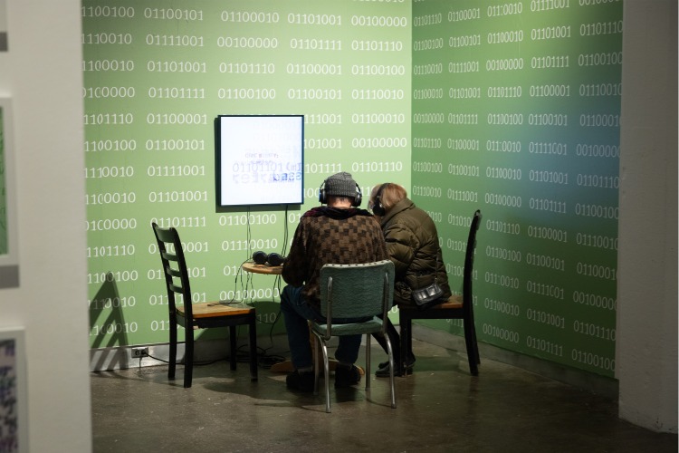 Installation shot of the Generation to Generation exhibition from C21's Aesthetics, Art, & AI program series, featuring gallery guests seated at table with headphones one, interacting with multimedia artworks. Installation shot of the Generation to Generation exhibition from C21's Aesthetics, Art, & AI program series, featuring gallery guests seated at table with headphones one, interacting with multimedia artworks.