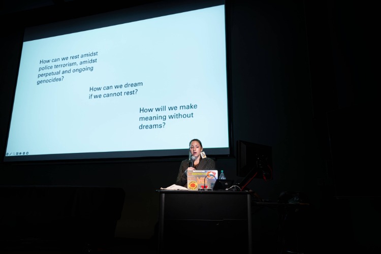 The artist Kite speaks at a podium in a darkened room, backlit by a giant projected slide, during her artist talk, part of C21's Aesthetics, Art, & AI program series. The artist Kite speaks at a podium in a darkened room, backlit by a giant projected slide, during her artist talk, part of C21's Aesthetics, Art, & AI program series.