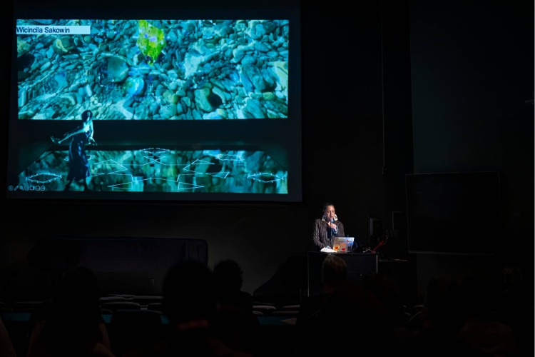 The artist Kite speaks at a podium in a darkened room, backlit by a giant projected slide, during her artist talk, part of C21's Aesthetics, Art, & AI program series. The artist Kite speaks at a podium in a darkened room, backlit by a giant projected slide, during her artist talk, part of C21's Aesthetics, Art, & AI program series.