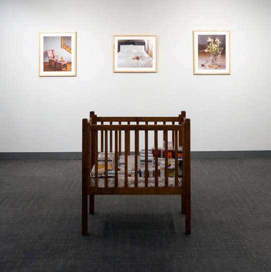 Image of an art installation featuring a crib,flanked by photographs mounted on a wall in the background